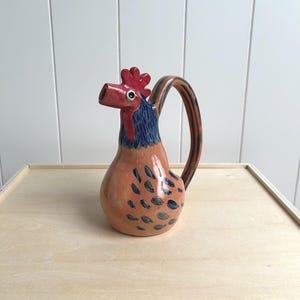 May include: A ceramic rooster pitcher with a red comb, beak, and handle. The body is peach-colored with blue teardrop-shaped markings. The neck is blue, and the handle has brown and orange stripes. The pitcher is on a light wood surface.