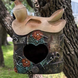 May include: A decorative birdhouse with a saddle-shaped roof and a heart-shaped opening. The birdhouse is painted in shades of brown, green, and orange, with floral accents and metal details. It hangs from a black wire, blending rustic charm with artistic design.