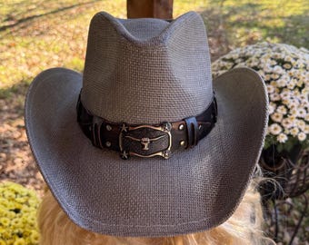 May include: A gray cowboy hat with a decorative leather band. The band features a metal buckle with a steer head design and star accents. The hat has a classic brim and a woven texture.