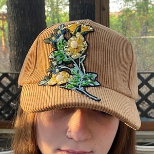 May include: A brown corduroy baseball cap with a floral embroidery featuring yellow, green, and blue flowers with pearl-like embellishments.