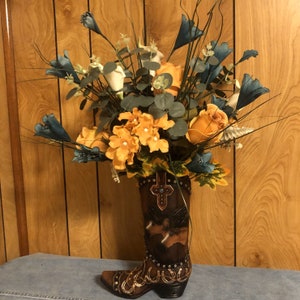 Hermoso jarrón marrón con forma de bota, con hermosos caballos y herraduras doradas. Arreglo floral de flores color melocotón y azul con alcatraces blancos.