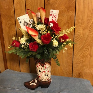Brauner Stiefel mit lustigen Spielkarten und Würfeln. Blumenarrangement aus roten und weißen Blüten mit viel Grün. Super für Diejenigen, die gerne Spiele spielen