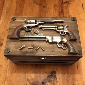 Può includere: Una scatola di legno con tre revolver antichi e proiettili. I revolver sono argentati e marroni, con dettagli intricati. La scatola è in legno scuro e presenta elementi metallici. Esposta su una superficie di legno.