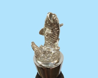 Pode incluir: Tampa de garrafa de prata com um design de peixe detalhado. O peixe está saltando para fora da água e tem um acabamento prateado.