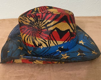 Puede incluir: Un sombrero de vaquero de paja con un diseño colorido. La corona presenta un patrón rojo, negro y dorado, mientras que el ala es azul con estrellas doradas. Una banda azul rodea la base de la corona.