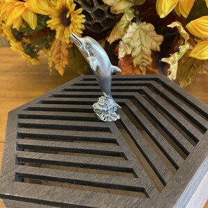 May include: A silver dolphin figurine leaping out of the water, perched on a textured base. The figurine is displayed on a dark wood hexagonal box with a geometric pattern.