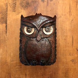 Peut inclure: Une figurine de hibou en bois brun avec de grands yeux détaillés. Le hibou a un motif sculpté sur ses plumes et une surface texturée.