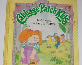 Op de afbeelding: Een gele boekomslag met een groen Cabbage Patch Kids-logo. De titel is "The Shyest 'Kid in the 'Patch". Het boek is een Parker Brothers Story Book met een verhaal van Mark Taylor en illustraties van Lynn Lace. De omslag toont een cartoonillustratie van een Cabbage Patch Kid-meisje met rood haar, een paarse jurk en een mand met bessen.