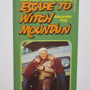 May include: A green book cover with the title "Escape to Witch Mountain" in orange letters. The book is based on the Walt Disney Studios' new movie and was written by Alexander Key. The cover features a photo of a man with two children.