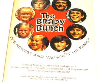 Op de afbeelding: Een boekomslag met een rode cirkel met de woorden "The Brady Bunch" in wit. De cirkel wordt omringd door acht kleinere cirkels, die elk een foto van een ander lid van de Brady-familie bevatten. De boekomslag bevat ook de tekst "TV's Newest and Wackiest Hit Family".