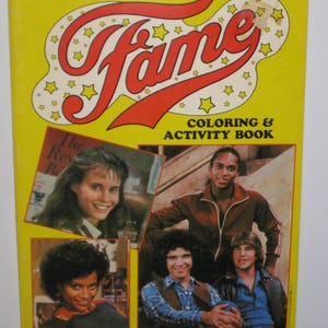 May include: A yellow coloring and activity book featuring the cast of the TV show "Fame". The cover features the show's title in red and white letters with a yellow background. The book includes images of the actors from the show, including a young woman with long brown hair, a man with short black hair, and two men with long brown hair.