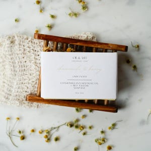 May include: A white soap bar labeled "Lavender & Honey" sits on a wooden soap dish, next to a natural fiber soap saver. The soap is unscented and contains chamomile-infused olive oil and beef tallow. Dried flowers are scattered around.