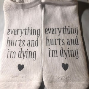 May include: A pair of white ankle socks with the text "everything hurts and i'm dying" printed on each sock. A small black heart is printed below the text on each sock.