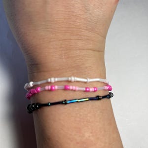 May include: Three beaded bracelets in white, pink, and black are stacked on a wrist. The top bracelet is white, the middle one is pink, and the bottom one is black with iridescent beads. The bracelets are delicate and simple.