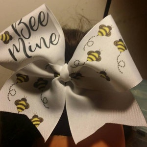 May include: White cheer bow with a black and white "Bee Mine" text and a pattern of yellow and black bees flying on a white background.