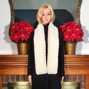 May include: A white faux fur scarf with a long, rectangular shape. The scarf is draped around the neck of a mannequin wearing a black dress. The mannequin is standing in front of a fireplace with red poinsettia plants on either side.