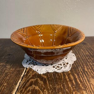 May include: A brown ceramic bowl with a decorative design. The bowl has a wide rim and a rounded bottom. It is sitting on a white doily.