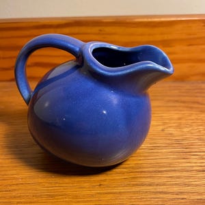 May include: A small, cobalt blue ceramic pitcher with a rounded body and a curved handle. The pitcher has a spout for easy pouring and sits on a wooden surface. The glossy finish reflects light, highlighting its smooth texture. This item is suitable for serving liquids.