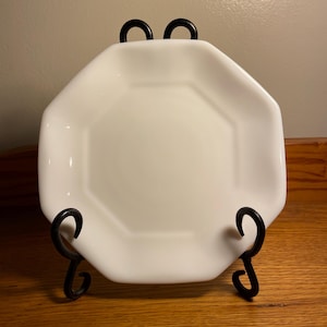 May include: A white, octagonal plate displayed on a black, wrought-iron plate stand. The plate has a glossy finish and is centered in the frame. The stand has decorative scrollwork.