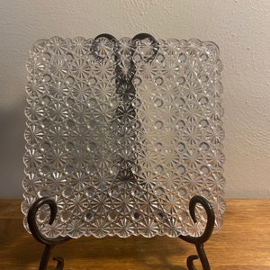 May include: Two clear glass square plates with a raised floral pattern, displayed on a black metal stand.