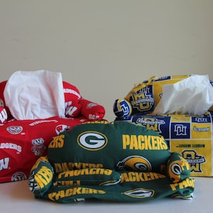 May include: Three decorative tissue box covers shaped like miniature sofas. One is red with white logos, another is green with yellow logos, and the third is blue and yellow with white logos. Each holds a stack of white tissues.