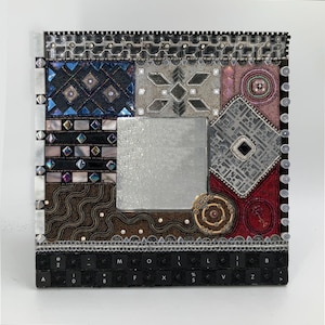May include: A square mirror with a mosaic frame made of various materials, including glass, metal, and beads. The frame features a geometric pattern with shades of blue, silver, red, and brown. The mirror is surrounded by a black keyboard with white keys.