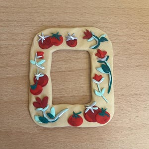 May include: A beige clay picture frame decorated with red, green, and white clay tomatoes and flowers.