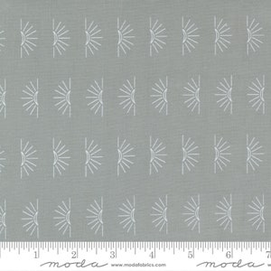 May include: Gray fabric with a white repeating pattern of stylized suns. The fabric is marked with a ruler in inches and the words "moda" and "www.modafabrics.com".