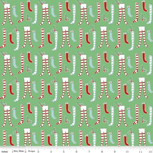 May include: A repeating pattern of Christmas stockings and legs on a green background. The stockings are red and white striped, solid red, and light blue. Some legs have holly sprigs. The design is by Riley Blake Designs.