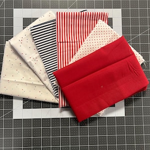 May include: A collection of fabric swatches in various patterns and colors, including red stripes, navy blue stripes, white with red dots, and solid red. The fabrics are arranged on a gray cutting mat with measurements.