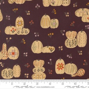 May include: A dark brown fabric featuring a repeating pattern of cream-colored pumpkins decorated with various geometric, floral, and star motifs. A white ruler at the bottom shows measurements in inches, confirming the scale of the print.