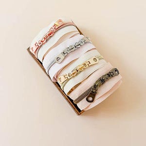 May include: A collection of four coiled zippers in shades of white and light pink, each featuring a distinct metallic pull tab in rose gold, silver, gold, and antique bronze finishes, arranged neatly within a small rectangular wooden tray.