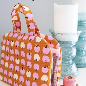 May include: A quilted orange fabric bag with a repeating pattern of pink and white apples. The bag has a clear plastic side and a handle. The text "THE TRUVY BAG" is at the top. The bag is from Knot + Thread Design.