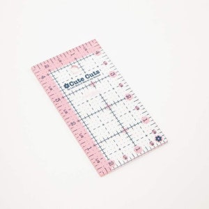 May include: A clear plastic ruler with pink markings and a grid pattern. It has a hole in the top and the text "Cute Cuts by Levi Mall of Bes in my Rennes" printed on it.