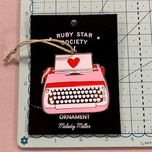 May include: Pink and gold typewriter ornament with a red heart on the paper. The ornament is on a black card with the text "RUBY STAR SOCIETY" and "ORNAMENT Melody Miller".