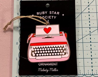 May include: Pink and gold typewriter ornament with a red heart on the paper. The ornament is on a black card with the text "RUBY STAR SOCIETY" and "ORNAMENT Melody Miller".