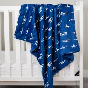 May include: A navy blue baby blanket with a white airplane pattern. The soft, plush blanket is draped over a white crib. The blanket features a repeating design of vintage biplanes.