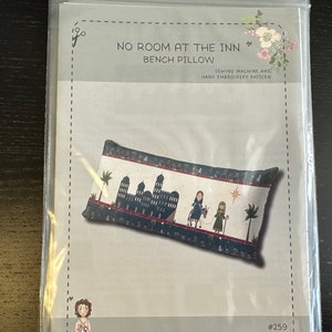 May include: A bench pillow pattern titled "No Room at the Inn." The pillow design features a nativity scene with a building, figures, and palm trees. The pattern is for sewing machine and hand embroidery.