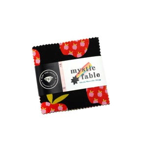 May include: A stack of fabric squares with a black background and a repeating pattern of red apples with pink centers. The fabric is wrapped with a white label that reads "mystic fable" and a black label with text and a diamond logo.