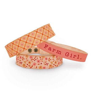 May include: Three decorative bracelets with floral and geometric patterns. One bracelet has the text "Farm Girl." The bracelets are in shades of orange, pink, and cream, with metal snap closures. The bracelets are likely made of fabric.