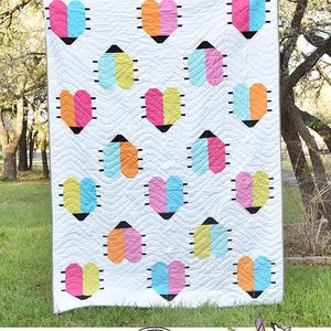May include: A white quilt with a repeating pattern of colorful heart-shaped designs. Each heart is divided into two colors, such as pink and blue, or yellow and orange. The quilt has the text "Love Bugs" and "Running Doe Quilts" on it.