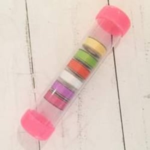 May include: A clear plastic tube with pink end caps containing several rolls of colorful washi tape. The tape rolls are yellow, green, orange, white, purple, and pink with striped patterns. The tube is on a white wooden surface.