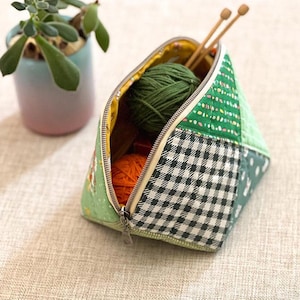 May include: A patchwork fabric knitting project bag with a zipper, containing green and orange yarn and wooden knitting needles. The bag features a mix of green, white, and black patterned fabrics. A small potted succulent is in the background.