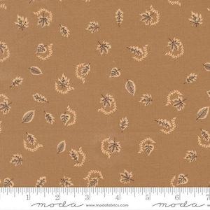 May include: A tan fabric featuring a repeating pattern of small dark brown leaves and light beige curved vine accents. A white ruler is visible at the bottom edge, showing measurements in inches, with the text moda www.modafabrics.com.