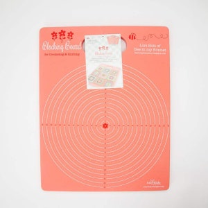 May include: A pink circular blocking board with white concentric circles and red flower accents. The board is labeled "Blocking Board for Crocheting & Knitting".