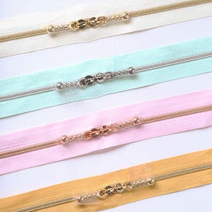 May include: Four fabric zippers arranged horizontally. The top zipper is white with gold hardware, the second is mint green with silver hardware, the third is light pink with rose gold hardware, and the bottom is mustard yellow with gold hardware.