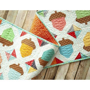 May include: A white quilt with a brown, green, blue, orange, and red acorn pattern. The quilt is folded in half and the fabric has a patchwork design.