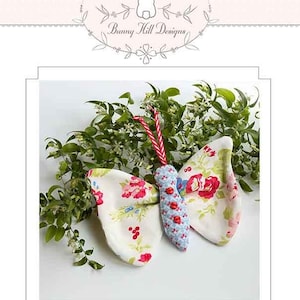 May include: A white fabric butterfly with red and blue floral patterns, hanging from a red and white striped string. The butterfly is surrounded by green leaves.