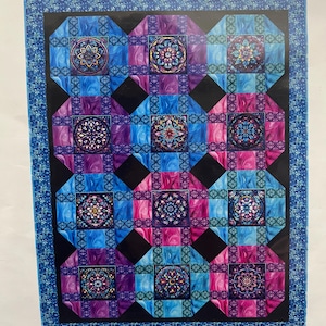 May include: Royale digital quilt top with a repeating pattern of squares and diamonds in shades of blue, purple, and pink. The quilt measures 57 inches by 73 inches. Each square features a floral design. The quilt is designed by Reed Johnson for Blue Bear Quilts.