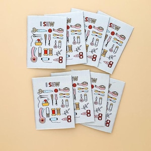May include: A collection of rectangular fabric labels with the text "I SEW" and "SEW" printed on them. Each label features colorful illustrations of sewing tools like scissors, needles, and measuring tape. The labels are arranged on a light beige surface.
